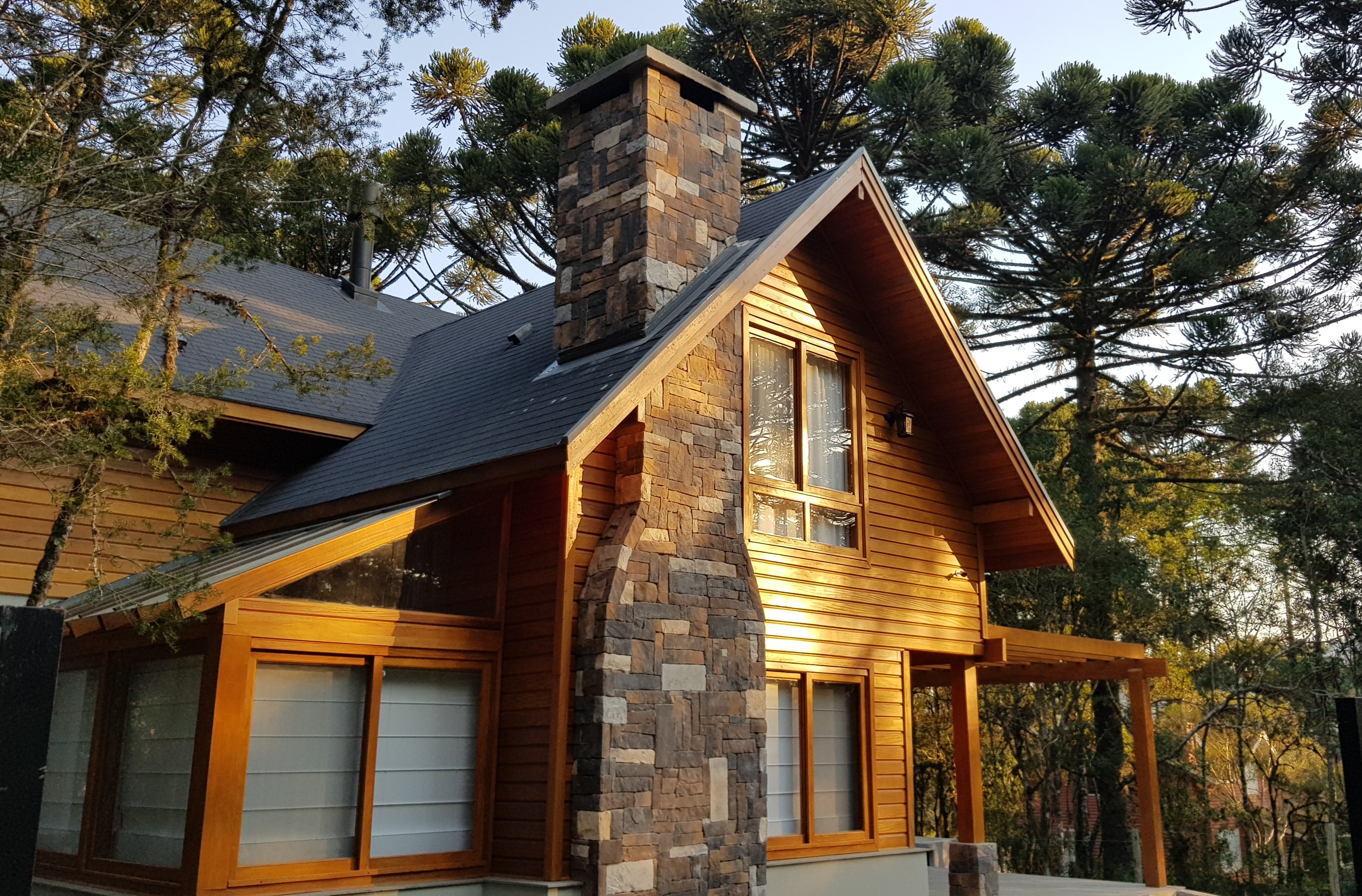 Chalé em madeira com chaminé de pedra entre araucárias, ao entardecer